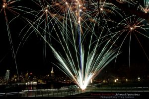 A silver fan cake firework display going off on a pier on the Hudson River with New York City in the background.